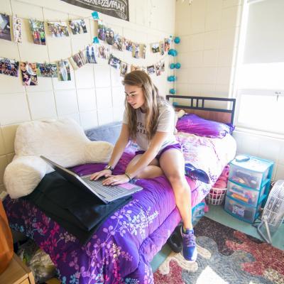 A young female Springfield College student sits on her bed in her residence hall while working on her laptop.